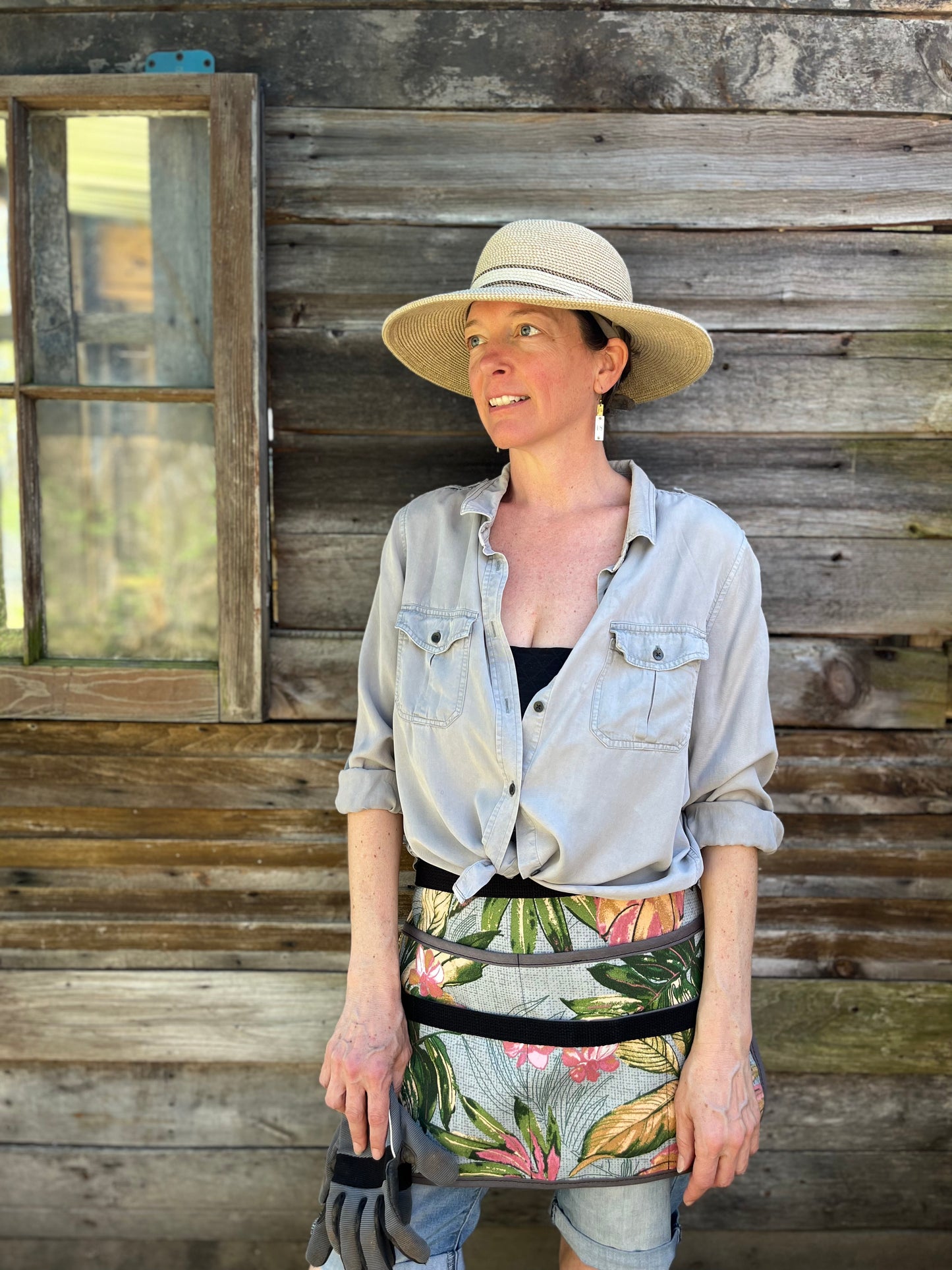 A woman in a printed half apron, sun hat and button down shirt stands in front of a barn, in the shade, and looks off into the distance, as though admiring her work in the garden.
